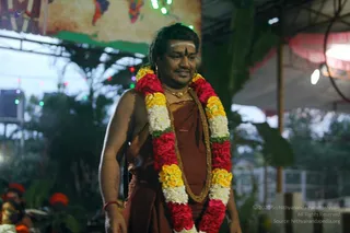 A man in a rust-brown dhoti-style garment with a bare chest and a long multicolour garland of red roses, yellow marigolds and white flowers draped over his shoulders and chest walks forward at an outdoor evening event. He wears a gold chain necklace, a gold-coloured thread or cord and gold earrings, and has a white tilak with a red bindi on his forehead. Behind him are strings of coloured lights, green foliage, a canopy, and orange flowers and an out-of-focus person at the lower left. The appearance of the man with garland and tilak is consistent with the page's a person reference but is not clearly distinctive enough to confirm, so he is described by role as a devotee-style figure in ceremonial dress.