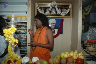 A shop interior during an inauguration. A man in an orange sleeveless robe with long swept-back dark hair and a rudraksha mala stands at centre, one index finger raised as he speaks. He appears to be a sannyasi in saffron. At left, a person in white holds a silver oil lamp with burning flames wrapped in yellow marigold garland. The counter in front holds coconuts, bananas, apples and other fruit. Shelves of packaged goods line the left wall, and a basket of red flowers sits at right.