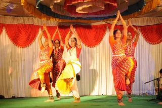 A photograph of a Bharatanatyam-style dance performance on a green stage under a draped canopy of red, blue and gold fabric with red and white swags behind. Five female dancers in colourful silk costumes with gold borders and ankle bells stand in a row with hands raised in anjali mudra above their heads, one lifting a leg in a dance pose. The central dancer wears a white and yellow skirt with a red blouse and gold necklace, and the others wear pink, orange and gold patterned costumes. A microphone stand is visible at the right edge. No a person candidate is visible.