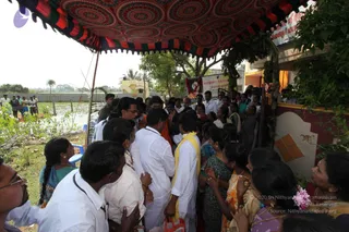 A crowd of devotees gathers outdoors beneath a red, green and cream patterned canopy supported by a pole. Men in white shirts and dhotis stand close together, one in a yellow shawl and another in orange clothing, pressing toward a doorway with pink and orange tiled walls. Women in saris and salwars, some with flowers in their hair, stand at the right with hands raised in reverence. A banner with a portrait and Tamil text is visible behind the crowd, near a water-filled field and palm trees.