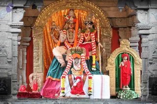 A photograph of a temple sanctum showing a large deity murti group framed by a gilded arch and orange-gold drapery. Two tall seated deity figures with ornate crowns, garlands of white and orange flowers, gold necklaces and red garments dominate the scene; one holds a long staff. In front, a smaller red-clad figure sits on a white cloth-draped pedestal beneath a garland, with a kalasha-like offering placed above it. On the right, a standing red-robed sage statue with a red turban and gold necklace stands in a green niche with a gold arch. Grey carved stone pillars frame the shrine.