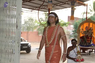A photograph of a procession at an ashram courtyard under a corrugated roof. A man with shoulder-length swept-back hair, a white forehead band and red dot, wears a saffron-orange cloth draped over his body with gold-bordered trim, a multi-strand ornate necklace with gold beads and rudraksha-style beads, and bangles on his arms and wrist, and walks forward through an open metal lattice gate. Behind him, a younger man in white dhoti and shawl with a white forehead band and beaded mala carries a brass tray with flowers and a lit oil lamp. To the right, a decorated palanquin or ratha draped in marigold garlands holds a deity murti. A white vehicle is parked outside and a brick wall and palm trees stand behind.