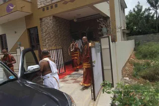 A photograph of a residential entrance with a tiled porch and a white metal gate. A bald man in saffron-orange dhoti and shawl, wearing rudraksha beads, walks through the gate onto a red-carpeted path, and a second saffron-clad man with a white flower garland stands beside him. Behind them, another person in a white shirt stands on the porch. In the foreground, a shirtless man in white dhoti with an orange cloth over his shoulder stands beside a dark car. A third saffron-clad figure stands at left. The a person candidate is the bald man in saffron who walks through the gate, shown with rudraksha beads and saffron clothing.
