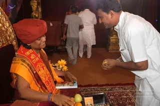 A woman seated on a gold-framed throne-like chair wears a red turban and an orange sari with a red sash printed with 'Jai Shri Ram' style Sanskrit lettering, and smiles while holding a small book. In front of her a man in a white kurta offers a yellow fruit, with fruits, yellow flowers and a small framed picture on a dark tray. Behind them two men walk across a room with red carpet and gilded decor. The setting appears to be an ashram hall.