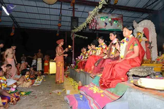 A night-time ritual in an open-sided hall with a corrugated metal roof. A priest in an orange turban and orange dhoti stands centre-left, holding a lit lamp toward a row of women seated on a raised platform. The women wear red and orange saris and garlands of yellow and white flowers. Behind them, a white and red decorated shrine holds a deity murti, with brass oil lamps in front. Men in white dhotis and bare-chested devotees sit or stand on the left, and yellow flower petals are scattered on the floor. A world map display hangs on the back wall, and hanging garlands decorate the ceiling.