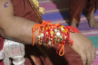 Close photograph of a man's forearm wrapped with several red and yellow rakhi threads, each with small gold and silver charms, beads and a metallic medallion. The arm rests over a maroon garment and a silver ornamental belt, while a striped colourful mat and bare feet appear at the lower right. No face is visible. The wrists show multiple rakhis, consistent with a Raksha Bandhan thread-tying moment.