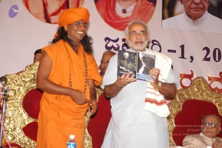 A photograph on a stage in front of a banner with Kannada script reading 'ಜನವರಿ 1, 2007' (January 1, 2007) and red lettering. A man with long dark hair wearing a saffron-orange robe and saffron turban, with a long rudraksha mala, stands at left smiling. Beside him, a man with white hair and beard in a white kurta with a white and red-bordered shawl draped over his shoulder holds up a book with a dark cover showing black-and-white portraits. Gold-carved thrones with red cushions flank them, and a seated elderly man in glasses is at lower right. Microphones and a water bottle are on the stage. A small portrait photo appears at top right. The orange-robed figure is an appearance-based probable Swamiji candidate, identified by the long hair, saffron turban and rudraksha mala cues.