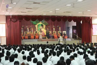 Photograph of a large hall filled with many seated people dressed in white, viewed from behind, facing a raised stage. On the stage, a central golden arched shrine holds a deity murti, with a green leafy backdrop above. A seated figure in a red and gold ornate chair sits at the centre, flanked by people in orange and saffron robes seated along the stage. A standing figure in white robes with hands folded and another in dark attire stand near the front of the stage. A cameraman with a tripod-mounted camera films from the front left of the stage area. Red drapes frame the stage, and ceiling fans and fluorescent lights hang overhead.