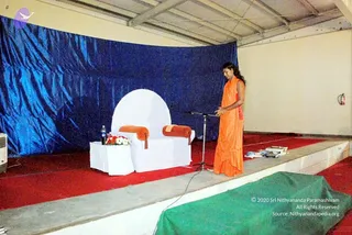 A stage set with a blue draped backdrop and red carpet. A white-covered armchair with orange cushions stands center left beside a white side table holding a water bottle and a red and white flower arrangement. A standing person in an orange saffron-coloured robe with long dark hair speaks at a microphone stand on the right of the stage, with a stack of books on the floor nearby. A green cloth covers part of the lower stage edge and a fire extinguisher is mounted on the wall at right. A logo appears in the top left corner.