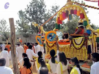 A festival procession on a dirt path with a decorated ratha, a chariot-like palanquin covered in yellow and orange marigold garlands, a tiered canopy with pink, green, orange and yellow tassels, and round blue-and-orange emblems on its sides. A figure in orange robes with a gold crown and garland sits on a raised seat at the top of the chariot. Around it, people in white dhotis and white kurtas walk, and several women in yellow-bordered white saris and orange sarees pull or steady the ratha with ropes. Men in orange and white stand in the background amid trees and a low wall.