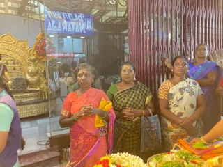 A photograph of several women standing with hands folded in a devotional posture near a glass-fronted display. Foreground, left to right: a woman in a coral-orange sari with a yellow cloth draped over her arm, a woman in a green blouse and dark plaid sari holding a grey-blue tote bag, and a woman in a floral white sari with yellow border. Further right, a woman in a blue-purple sari stands beside a red metal shutter. In the lower foreground, baskets of white jasmine garlands, marigolds and green leaves are arranged. Behind the glass, a golden throne with lion-like carvings and a seated golden figure are visible, with a blue signboard reading Lalitha Natraja above. Four women are visible.