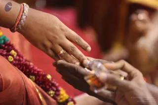 Close-up photograph of two hands meeting in a ritual exchange. On the left, a woman's hand extends forward, wearing a thin gold ring on the ring finger and a red and grey thread bracelet on the wrist, with a sleeve of rust-red fabric and a red and yellow flower garland beside it. On the right, a darker hand cups beneath with a small glass vessel holding a golden liquid, possibly oil or ghee. The background is blurred warm red and gold tones.