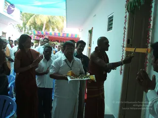 A corridor opening ceremony in a white-walled building with a striped canopy outside. A bald priest in a red-gold dhoti with a garland and sandalwood-marked chest cuts a yellow ribbon with scissors at a doorway decorated with red flower garlands. A man in white dhoti and shirt holds a tray of flowers and offerings, and a woman in a red sari stands to the left. Several onlookers in white and dark clothes stand behind, with some clapping and others with hands folded in greeting. Blue plastic chairs line the corridor.