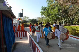 A photograph of a procession on a concrete path beside a small white temple building with a blue cloth hanging at its entrance. In the foreground, several men carry drums: a bearded man in a blue t-shirt and dark dhoti plays a drum held at his side, while men in white shirts and white dhotis, one with a pink sash, play drums and cymbals. Behind them, a group of people in saffron/orange robes walk along the path carrying long poles and a striped yellow-and-red parasol. A woman in an orange sari with a garland stands among them. A water tank sits on a pole overhead, with trees and greenery behind. Other men in white dhotis and shirts walk in the background.