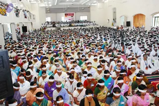 A large indoor hall filled with hundreds of seated devotees, most wearing white blindfolds tied across their eyes. Women in saris of many colours, including purple, teal, red, pink and yellow, sit closely on the floor, with some men in white shirts among them. A banner with a photograph hangs on the back wall, and people are seated in tiers on a raised stage at the rear. A black speaker stands at the left edge, with video camera tripods visible on the right. Ceiling fans, arched doorways and white walls frame the scene.