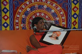 A man seated cross-legged on an orange cushioned couch in front of a red, yellow and blue patterned wall hanging with elephant and floral medallion motifs. He wears an orange-red robe with his bare arm visible and smiles while holding up a framed portrait. A microphone on a stand is in front of him. The framed portrait shows a smiling man with long dark hair, an orange turban and orange robe, labelled as a depiction. A small cloth-covered book rests on the couch at lower left.