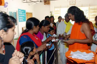 A photograph inside a bright hall with a wall clock and a blue information sign. A long-haired man in saffron-orange robes stands at right, handing small items or packets to a line of devotees. Several women and girls queue along a metal railing; one woman in a red shawl and blue blouse holds up small white papers, a girl in a blue top reaches forward, and a young woman in a pink top stands nearby. Behind them a man in white with a yellow scarf waits. An orange plate holds a stack of white papers at lower right. The man in orange has a long, swept-back hairstyle and a rudraksha-style mala visible at his chest, which is a visible appearance cue that matches a person, so the identification is probable. He appears to be distributing prasad or papers to the line.