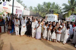 A large outdoor crowd stands barefoot on sandy ground in front of a white booth with banners and signage, with coconut palms and a thatched hut behind. A row of devotees in white clothes stands with palms pressed together in namaskar, including a man in white kurta and white dhoti, a man in black shirt and black dhoti, a woman in white with a mala, and several children in white in front. A woman in a blue-and-white sari stands at left and an older woman in white stands at right with hands folded. A white van labelled Be Blissful and Ananda Kailasam is parked behind them, with a speaker on top. Banners and posters are visible on the booth, one showing a photograph of a woman, and a map poster is mounted on the right. The scene suggests a devotional gathering or procession with people in prayer posture.