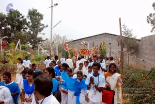 A crowd of devotees walks along a dirt path in a garden-lined compound, following a decorated procession vehicle. Young men and women in white shirts and white dhotis or saris, several wearing blue shawls, move forward with hands pressed together in namaste and rudraksha malas around their necks. In the centre background, an ornate orange and gold ratha or palanquin with a canopy and decorative umbrellas is carried along, flanked by green banana-leaf decorations and red flowers. A stone-walled building with a metal-framed roof stands behind, with street lamps and a wire fence to the right. A bald man in white stands among the crowd on the left.
