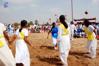 Women in white salwar-style outfits with yellow sashes play a game on a sandy ground in an outdoor open field. Several women wear yellow sashes over white clothes, one wears a blue top over white, and a woman in a white shirt stands at the right edge. A large crowd of people in white and some in blue, red and green clothing sits and stands behind a rope boundary. A tent with striped canopy stands at the right, orange flags flutter on poles, and a clay pot hangs from a rope near a tall pole. Palm trees and a hazy sky form the background.