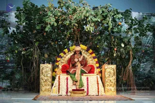 A seated figure sits on an ornate gilded throne backed by a garland of marigolds in yellow, orange and purple, set before a stage backdrop of leafy branches with hanging orange fruit. The figure wears a rust-brown tunic, a green sarong, a cream shawl and a cream turban, with hands pressed together in anjali mudra. The throne is decorated with white and orange flower garlands and a red cushion, and a small pedestal of flowers sits at the base. Gilded side panels frame the seat. The seated figure is an appearance-based probable candidate given the throne, turban and shawl, but the cues are not distinctive enough to confirm identity, so the figure is described by role as a seated person on a throne.