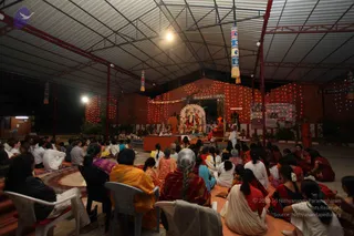 A night-time photograph inside an open-sided temple hall with a corrugated metal roof, red-painted pillars and strings of red fairy lights on the walls. Dozens of devotees sit on the floor and on white plastic chairs facing a raised shrine at the far end, where a large deity murti with a halo-like backdrop stands beside other figures and a tall orange-robed figure in a white shawl stands at the side. Many women wear red, orange and pink saris, and a few men wear white. Signboards on the walls, a sign reading Sri Nithyananda Paramashivam Temple, and hanging decorative banners complete the scene.