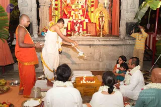 A homa ritual at a temple shrine with a deity murti decorated with red and yellow garlands behind an altar. A priest in white dhoti with gold trim pours a liquid from a bottle into a square fire pit with a flame. To the left, a shaved-headed man in orange robes and a white garland stands holding a metal bowl. Seated devotees in white clothes with flower garlands sit around the fire pit, including a woman in an orange sari holding a small child in teal, and a man in white. A videographer in a striped yellow shirt films on the right. Brass vessels, bananas and a bowl of rice sit on the floor.
