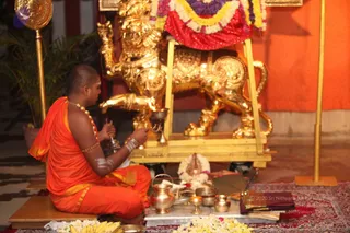 A photograph of a priest seated cross-legged on a low wooden platform performing puja before a large gilded lion-form vahana with a red and purple floral garland draped over its back. The priest, shaved-headed and bare-chested, wears an orange dhoti with gold border, a gold necklace and gold armbands, with white sacred markings on his arm. He holds a small bell and a ritual implement. In front of him stand brass vessels, a small kalasha-style pot, flower garlands, bananas and a small deity murti on a white marble plate. Orange drapery and a potted palm frame the background, with gold standing poles on either side.