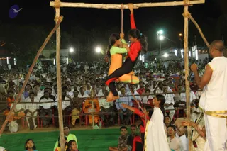 Night outdoor event on a green mat with a wooden frame. Two girls in orange, green and red athletic outfits hang from a horizontal bar and rope, one holding the other while suspended in the air. A large seated crowd of hundreds watches from red plastic chairs behind a rope barrier, many men in white dhotis and shirts, some in saffron cloth. On the right, a man in white dhoti with gold border stands holding a microphone, and a woman in white sari with gold border watches nearby. Bright floodlights and stage banners are visible in the background.