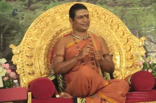 A man sits cross-legged on an ornate gold throne with a large gilded backrest and red cushions, in front of a backdrop of green foliage. He wears an orange-saffron garment with a yellow edge, multiple rudraksha malas and beaded bangles on his arms, and a white-and-red forehead mark. His hands are raised in front of his chest in a teaching gesture. Pink and white flowers and a water bottle sit beside the throne. Given the page's indication that a person was present and the strong cues of saffron clothing, rudraksha malas and a throne, this is an appearance-based candidate for a person, seated as the teacher at a satsang.