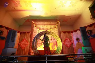 A performer stands on a raised stage dressed as a deity with a trident, wearing a green and gold costume, a garland of flowers and beaded necklaces, and dark skin paint. The performer holds a long trident in one hand and stands before a large golden circular frame with a glowing fire and a flame-lit backdrop. A marigold garland arches over the frame. Red kolam-style diamond panels with white patterns, red and white draped curtains, floral decorations and two cooler units frame the stage under a decorated ceiling with floral motifs. The scene resembles a theatrical or festival tableau. No a person candidate is visibly identifiable from appearance, so no in-person presence is claimed.