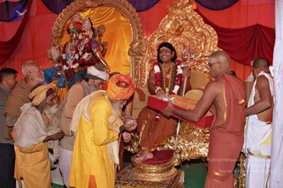 A photograph inside a red and gold tent at a festival camp. A seated figure on an ornate gold throne wears an orange robe, a white and pink flower garland, gold necklaces and has long dark hair. Seated figure holds a brass trident. Before the throne, several elderly sadhus in saffron, yellow and white clothing and turbans bow and offer things, one holding a small packet. A bald priest in an orange garment and white-clad attendants stand nearby holding a red box. Behind them stands a painted or sculpted deity murti with a crown, garland and mace. Red and purple drapes frame the scene. a person is seated on the throne wearing rudraksha and gold necklaces with flowers.