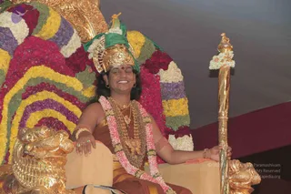 A smiling seated figure sits on an ornate golden throne (peetha) decorated with a large arch of layered flower garlands in red, yellow, purple and white. The figure wears a tall golden crown with a green cloth and a red bindi, multiple gold necklaces and a pink-and-white flower garland, bangles on the wrists, and an orange-and-gold garment. Beside the throne stands a tall golden staff topped with a small figure and a white flower garland. The throne's armrests end in carved golden lion heads. The figure appears to be a presented or honoured person seated on a decorated throne; Swamiji (probable) cues are the swept-back hair, crown, saffron-orange clothing and gold necklaces.