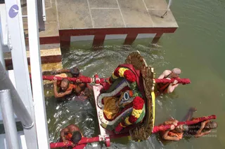 A decorated wooden palanquin-style deity carriage with a gold-framed rim sits on a red-trimmed platform in green water, garlanded with red roses, yellow and white flowers and a dark red cloth. A golden-yellow cloth drapes the figure within. Several men in orange and saffron garments, some with shaved heads, wade in the water pulling the carriage on thick red ropes, while one man on the right upper side holds a rope. Stone steps and a water spout appear at the top of the frame.