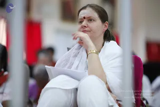 A woman with her hair pulled back sits in a white outfit with a white shawl draped over her shoulders, a gold bangle on her wrist and a red dot and white forehead marking. She rests one hand near her face and holds a stack of white papers on her lap, looking off to the side. Blurred figures in white and saffron sit in the background, with red fabric and metal pillars in the foreground.