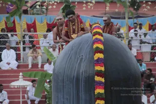 Outdoor temple-style ceremony at a large dark polished black stone lingam covered with a garland of yellow, orange, red and purple flowers. A central figure in a red garland and gold necklaces with a rudraksha mala, forehead marked with white tripundra, rests his hands on the top of the lingam while bending over it, flanked by two shaved-headed priests in saffron-brown cloth with white forehead marks and beaded necklaces. Behind them a crowd of people in white garments sits on red-stepped seating beside a fence; banners in red, yellow and blue hang along the back, with a banana-leaf plant and a white ceremonial umbrella in the foreground.