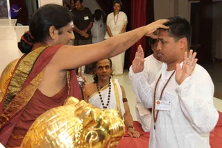 A photograph of a blessing ceremony indoors. A woman with hair tied back, wearing a maroon and gold-bordered wrap, places her right hand on the head of a young man in a white kurta with a rudraksha mala and a name badge, while he holds his palms up. Beside her, a man in white with a rudraksha mala and gold-bordered cloth sits watching, and a golden kalasha-like vessel with a gold holder sits in the foreground near a small water vessel. Behind them, a man in a dark shirt with a badge and a woman in a white sari with a rudraksha mala stand in the background with a red curtain. Scattered visible dots of sacred marks and gold ornaments are present.