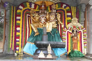 A photograph of a temple shrine with golden deity murti seated on a black peetha around a dark shivalinga, dressed in teal-green draped cloth. The two gold murti wear red-and-gold turbans, jewelled necklaces and earrings and hold ceremonial staffs. Behind them is a wall of marigold and mixed-colour flower garlands in yellow, orange, red, purple and white. On the right, a smaller wooden standing figure in saffron-brown garments with a rudraksha-style necklace stands in an arched frame of flowers. Small white and gold figures sit at the base. No living people are visible.