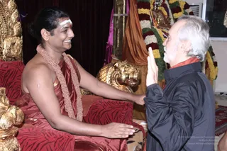 A photograph of two men facing each other in a richly decorated room with gold lion-head carvings and red tiger-striped cushions. On the left, a smiling man with a swept-back dark hairstyle, a white tilak on his forehead, a red-orange silk garment and a beaded rudraksha-style necklace sits on a red throne with his hand extended toward the other man. On the right, an older man with white hair and a white beard, wearing a dark jacket over a red-orange garment, raises his palm toward the seated man. Behind them hang a garland of orange, red and white flowers, a gold statue, and orange draped cloth.