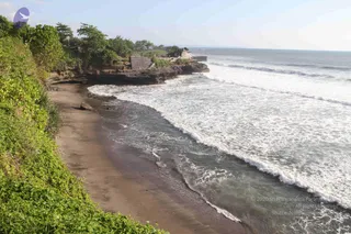 A photograph of a dark volcanic-sand beach on a tropical coastline, with white surf rolling in from the right and green vegetation covering the cliff on the left. On a rocky headland in the background, a small stone temple structure with a dark tiled roof sits among trees, consistent with a Balinese sea temple. No people are visible; the scene shows only the shoreline, waves and foliage.