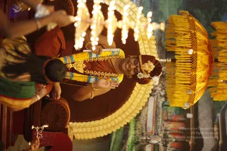A photograph, rotated sideways, of a seated figure on a dark brown throne with a gold scalloped edge, receiving a ceremonial ritual. The seated person wears a rust-red garment, a rust-red turban with a gold ornament, a multi-strand garland of yellow and blue flowers, a gold pendant necklace, gold bangles and a gold brooch on the turban. The person holds a long decorated staff tied with garland. In the foreground at left, a partially visible person in a green and gold garment holds up a lit ritual lamp cluster of flames. Behind the throne stand a large yellow fringed parasol and a painted backdrop of a garden scene. The figure is seated and the scene is a padapuja-style honouring with lamps.