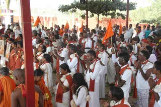 A photograph of a large outdoor gathering of devotees standing with palms pressed together in namaskar beneath an open pavilion with red pillars. Many men and women wear white dhotis or saris with red shoulder cloths, and some carry white bundled offerings. Several saffron-robed sannyasis stand in the foreground, one with a shaved head and visible rudraksha beads. Orange flags flutter in the background among trees and red walls, and a framed picture is held at the left edge. Several dozen people are visible in the scene.