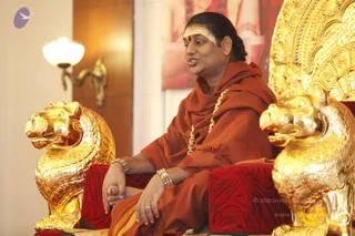 A seated man in a rust-orange robe and shawl gestures while speaking, sitting in an ornate gold throne with gilded makara-style lion-head arms and red tiger-striped cushions. He wears a long gold necklace, beaded bracelets and a white sacred ash marking with red bindi on his forehead. Behind him hangs a red-toned picture panel, and a brass wall sconce lights the cream wall at left. The scene resembles a seated satsang discourse.
