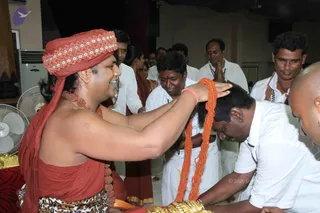 Indoor photo of a seated man in a red-orange turban with white floral trim and a rust-red shawl, wearing a red beaded necklace and rudraksha-style beads; his bare arm reaches forward to bless a kneeling man in a white shirt who bows his head. Behind them several people in white shirts and saffron-orange garments stand, one with palms pressed together in greeting, and a long orange-red beaded garland is draped over the bowed man's head. a person is a probable identification based on the saffron turban, red shawl and beaded adornments on the seated figure who is blessing the devotee.