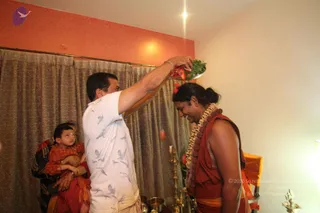 A man in a white short-sleeved shirt printed with grey bird motifs holds a silver plate of red flowers and green leaves high above the head of a smiling man with a long dark ponytail. That man wears an orange-red shawl and a large cream-and-brown flower garland, and his head is bowed. Behind them a woman in red holds a toddler in a red outfit with gold bangles. Peach-coloured walls, grey curtains, brass oil lamps and a lit lamp on the right suggest a home puja.