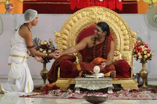 A seated man on an ornate gold-and-red throne, wearing a saffron-orange robe, rudraksha mala necklaces of beads, and a bare upper body, with white sacred markings on his forehead, smiling as he extends his hand. Before him, a young attendant kneels in white dhoti and shoulder cloth with a white head covering and rudraksha beads, offering or receiving his hand near a brass pot of flowers. Between them stands a silver tray with a white object and a small pot of flowers; red and white roses fill brass vases on both sides. Probable Swamiji cues: long swept-back hair, saffron robe, and rudraksha malas on a throne. The setting is a temple-like hall with red drapes and a red carpet, and a fan is visible at right. Possible padapuja or darshan ritual.