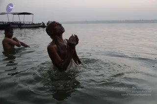 A photograph of a man standing waist-deep in a wide river at dawn, bare-chested with wet hair slicked back, palms pressed together in prayer above his head and face lifted skyward. A white cloth is wrapped around his lower body. To the left, another bare-chested man stands partly submerged, arms resting on the water. Behind them a covered wooden boat is moored near the shore under a pale, hazy sky. The scene suggests a ritual bath or water prayer.
