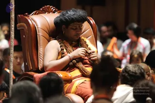 A seated man in a large brown leather armchair with carved wooden arms and a scrolled crest is the central subject, wearing an orange-brown robe with yellow-gold borders and a red-orange sash. He has a red bindi and a tilak mark on his forehead, a rudraksha mala of large beads draped across his chest, and a green bead strand at his neck, while his hands work with the beads of the mala and his eyes are lowered. Audience members in orange, white and dark clothing sit in the blurred foreground and background, and a metal stand pole is at the left edge. Appearance-based a person cues include the rudraksha malas and saffron-toned robe with a ceremonial gold border, but the identity is uncertain from appearance alone and the figure is a seated devotee-style presence in a throne-like chair.