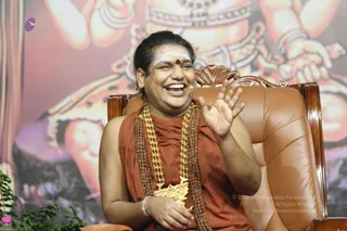 A smiling man with a shaved head and white vibhuti lines on his forehead, a red kumkum dot between his brows, sits on an ornate brown leather throne chair. He wears an orange-red saffron robe draped over one shoulder, a long gold necklace and a thick rudraksha mala across his chest. He raises one hand in an animated gesture while the other rests in his lap. Behind him is a large painted backdrop of a deity with multiple arms and bangles, and a small potted plant sits at the lower left. The scene appears to be a seated talk or Q&A session.