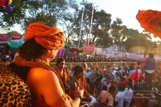 Outdoor photograph of a Kumbh Mela procession at dusk, with a crowd of devotees behind metal barricades. In the foreground a sadhu wearing a tightly wrapped orange turban and an orange-beaded rudraksha-style necklace faces away from the camera, a leopard-print cloth over his shoulder, one hand raised. Beside him a drummer in orange robes and beaded necklaces plays a drum, and another sadhu in an orange garment stands nearby. Red and orange flags and banners fill the background, with a cameraman on a raised platform at right. Festival trees and tents surround the scene.